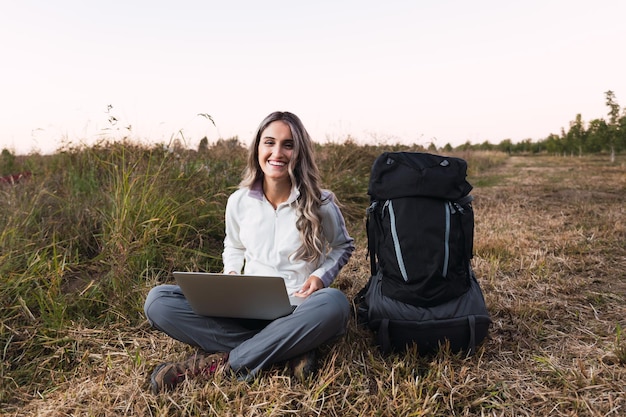 Mujer joven con una mochila a su lado usando una computadora portátil para teletrabajar en medio del campo