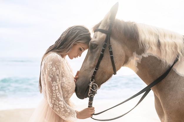 Mujer joven de moda feliz en un vestido blanco que presenta con un caballo en la playa.