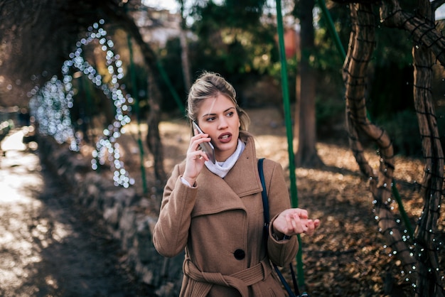 Mujer joven de moda que habla en el teléfono elegante en el parque de la ciudad.