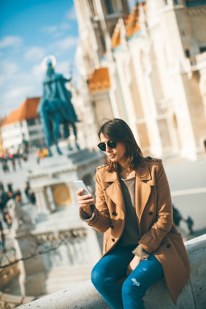 Mujer joven de moda que se sienta al aire libre y que usa el teléfono móvil