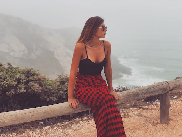 Mujer joven de moda sentada en la barandilla contra las montañas