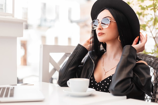 La mujer joven del modelo de moda en ropa hermosa elegante negra en gafas de sol está descansando sentado en la silla de la vendimia en el café al aire libre de verano en un día soleado. La muchacha atractiva tomó café. Buen fin de semana.