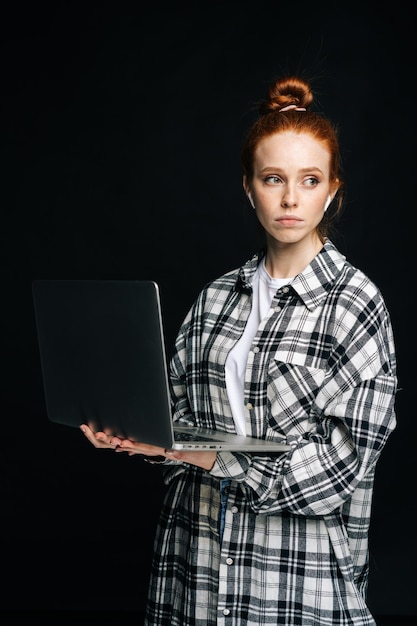 Mujer joven molesta sosteniendo una computadora portátil usando auriculares inalámbricos y mirando hacia otro lado