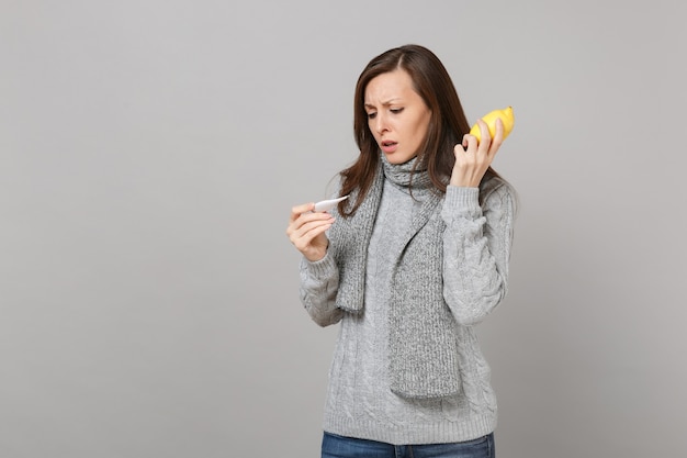 Mujer joven molesta en suéter gris, bufanda con limón, mirando el termómetro aislado sobre fondo de pared gris. Estilo de vida saludable, tratamiento de enfermedades enfermas, concepto de estación fría. Simulacros de espacio de copia.
