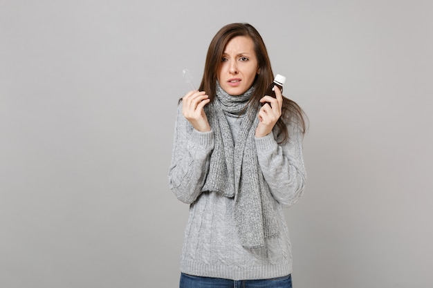 Mujer joven molesta en suéter gris, bufanda sosteniendo vierta medicina líquida y cuchara aislada sobre fondo de pared gris. Estilo de vida saludable tratamiento de enfermedades enfermas, concepto de estación fría. Simulacros de espacio de copia.