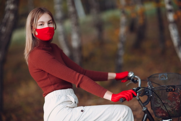 Mujer joven montando bicicleta en guantes rojos y mascarilla en el parque de otoño.