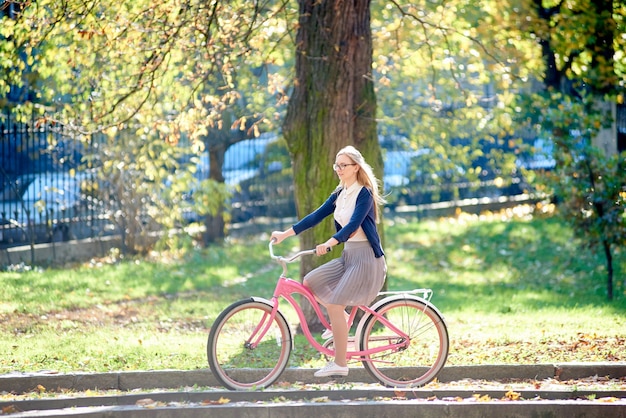 Mujer joven montando bicicleta Pink Lady a lo largo de la acera pavimentada en el parque en un día soleado de otoño