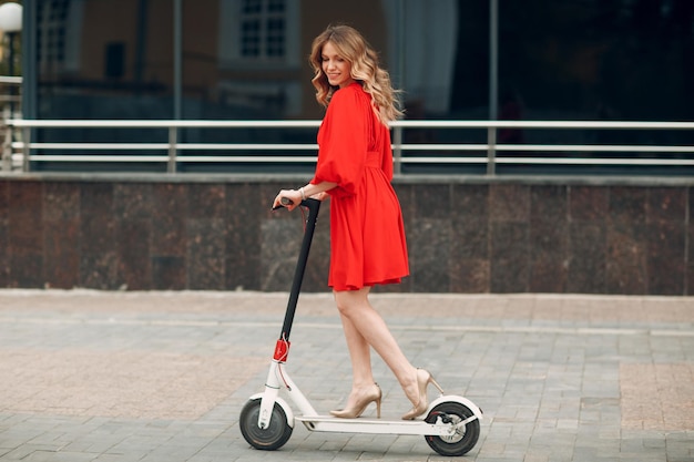 Mujer joven montando scooter eléctrico con vestido rojo en la ciudad