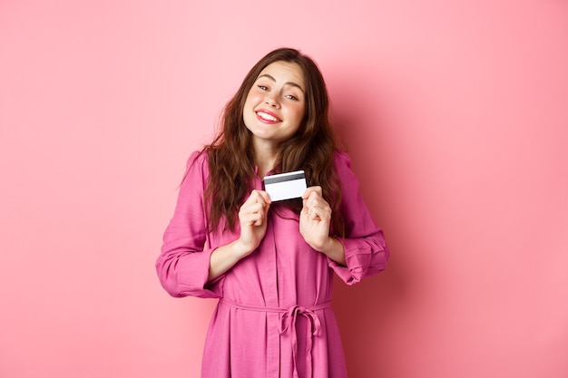 Mujer joven morena abrazando su tarjeta de crédito con deleite, sonriendo complacida, de pie contra la pared rosa. Copia espacio