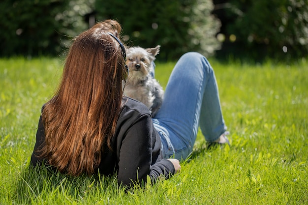 Mujer joven morena en auriculares relajándose en la naturaleza con mascota perro pequeño disfrutando de la vista, tumbado sobre el césped en el parque