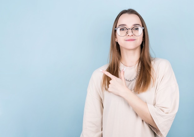 Mujer joven morena en una camiseta amarilla con una cadena alrededor de su cuello y gafas sobre un fondo azul apuntando