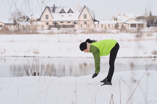 Mujer joven morena en chaqueta verde de pie en el lago y hacer ejercicio antes de correr