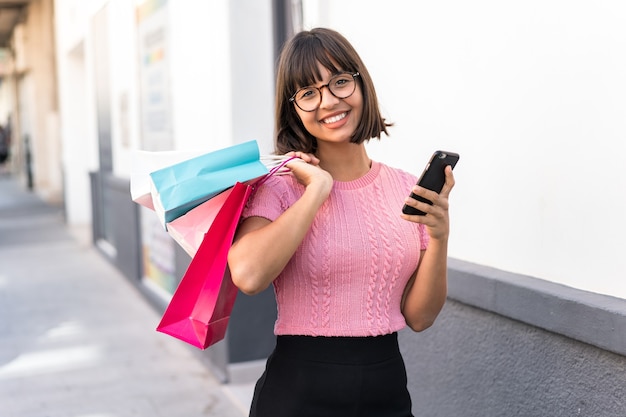 Foto mujer joven morena en la ciudad sosteniendo bolsas de la compra y escribiendo un mensaje con su teléfono celular a un amigo