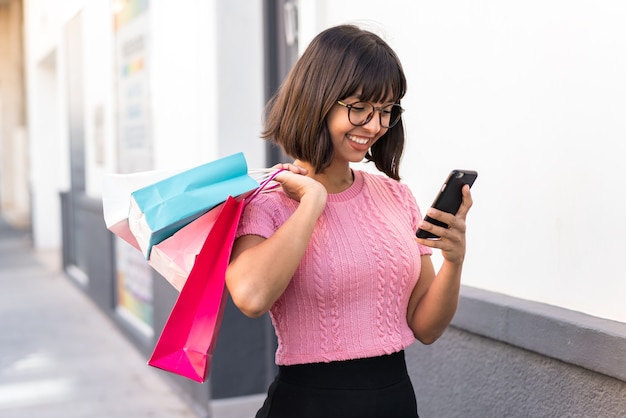 Foto mujer joven morena en la ciudad sosteniendo bolsas de la compra y escribiendo un mensaje con su teléfono celular a un amigo