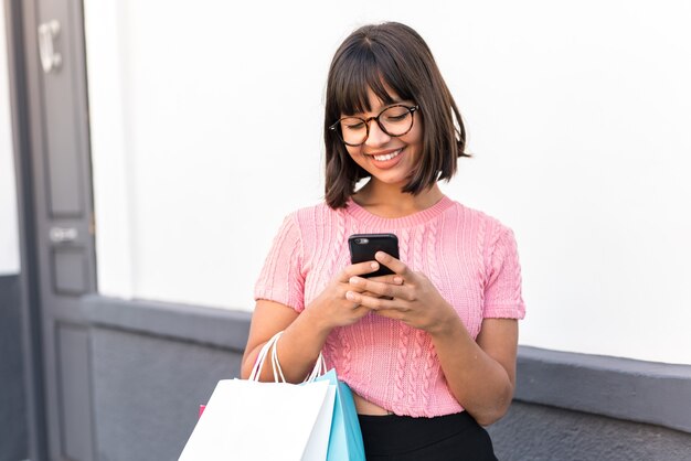 Foto mujer joven morena en la ciudad sosteniendo bolsas de la compra y escribiendo un mensaje con su teléfono celular a un amigo