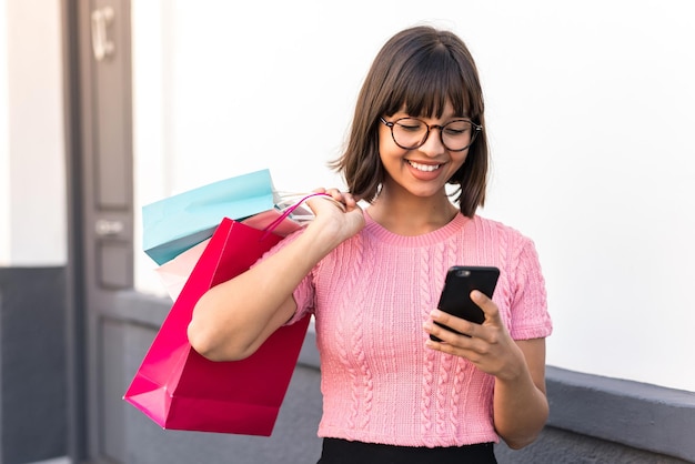 Foto mujer joven morena en la ciudad sosteniendo bolsas de la compra y escribiendo un mensaje con su teléfono celular a un amigo