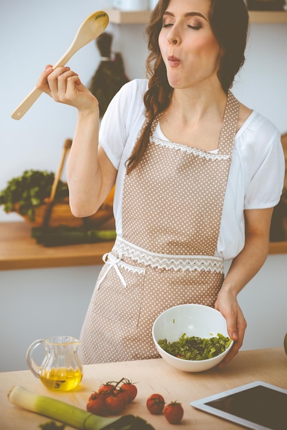 Mujer joven morena cocinando en la cocina. Ama de casa con cuchara de madera en la mano. Concepto de comida y salud.