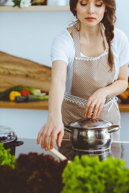 Mujer joven morena cocinando sopa en la cocina. Ama de casa con una cuchara de madera en la mano. Concepto de comida y salud.