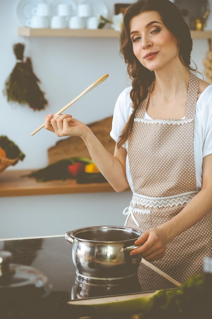 Mujer joven morena cocinando sopa en la cocina. Ama de casa con una cuchara de madera en la mano. Concepto de comida y salud.