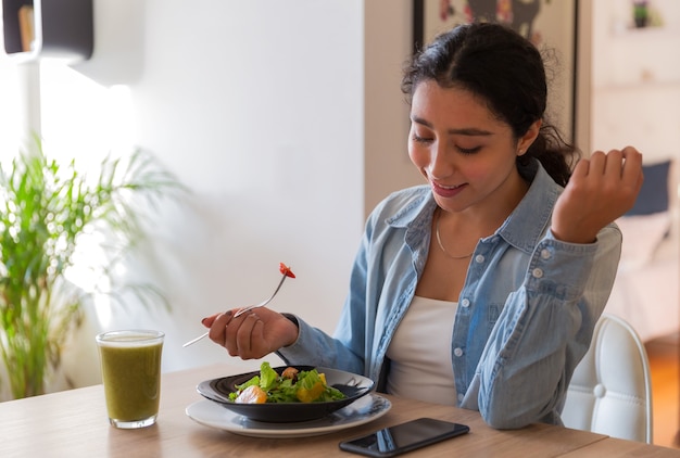 Foto mujer joven morena comiendo una ensalada en su comedor ella está usando su teléfono