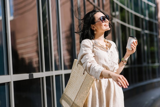 Mujer joven morena con gafas de sol y bolso con café caminando por la ciudad. Retrato de estilo de vida de mujer