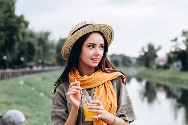 Mujer joven morena hipster beber jugo de naranja al aire libre, sonriendo, disfrutando de sus vacaciones. Vacaciones de verano en la ciudad. Closeup retrato de una niña en un elegante sombrero en el parque.