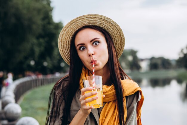 Mujer joven morena hipster beber jugo de naranja al aire libre, sonriendo, disfrutando de sus vacaciones. Vacaciones de verano en la ciudad. Closeup retrato de una niña en un elegante sombrero en el parque.