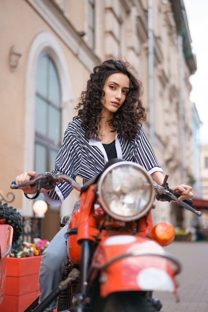 Mujer joven morena en jeans y camisa a rayas se sienta en una motocicleta roja al aire libre.