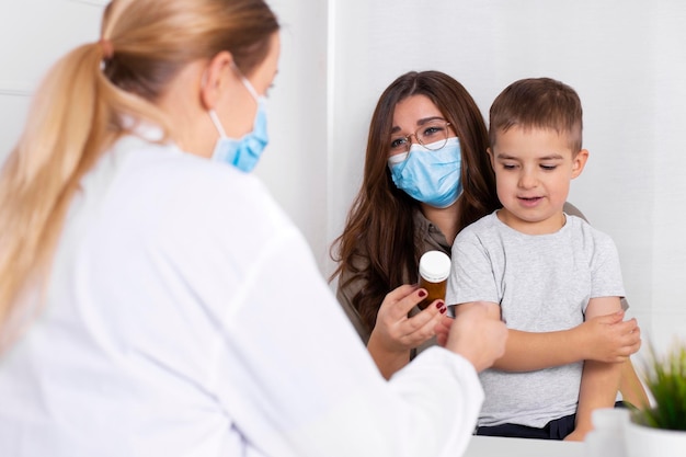 Mujer joven morena con niño tras consulta en el consultorio del pediatra. Médico, niño y madre con mascarillas durante el brote de coronavirus y gripe. Protección contra el virus. COVID-19.