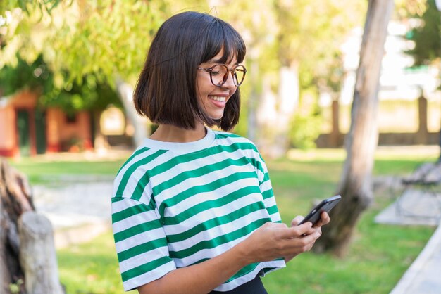Mujer joven morena en el parque enviando un mensaje o correo electrónico con el móvil