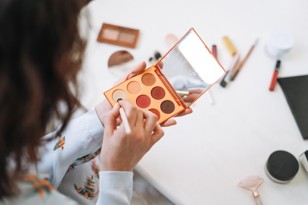 Mujer joven morena en pijama azul aplica maquillaje con paleta de escultura facial en las manos en casa