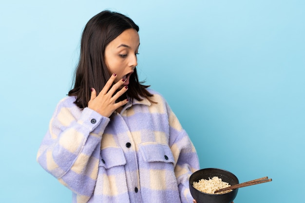 Mujer joven morena de raza mixta sobre pared azul con expresión facial sorprendida y conmocionada mientras sostiene un plato de fideos con palillos.