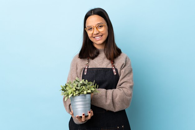 Mujer joven morena de raza mixta sosteniendo una planta sobre risa azul aislado.