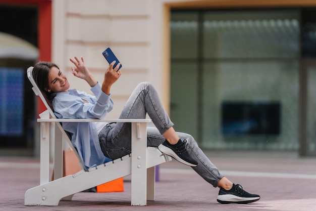 Mujer joven morena sentada afuera en una silla de madera haciendo una videollamada muestra el signo de ok Joven bloguera hace selfie sonriendo muestra el signo de ok en la cámara del teléfono Mujer alegre de moda en un descanso informal