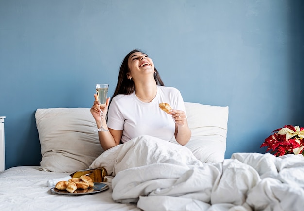 Mujer joven morena sentada despierta en la cama bebiendo champán comiendo croissants