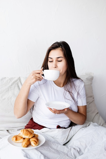 Mujer joven morena sentada despierta en la cama con globos en forma de corazón rojo y decoraciones bebiendo café comiendo croissants