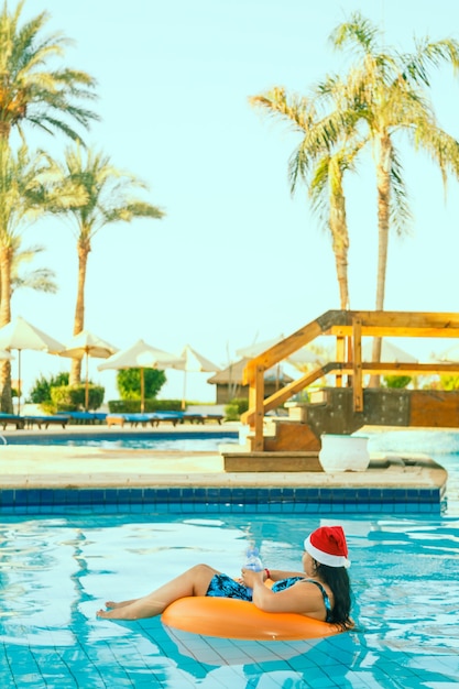 Mujer joven morena con sombrero de santa claus en un círculo de natación en la piscina durante las vacaciones de Navidad.