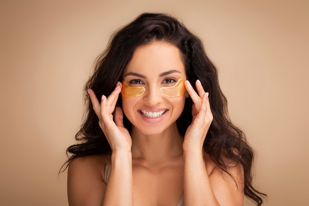 Mujer joven morena sonriente aplicando parches dorados debajo de los ojos