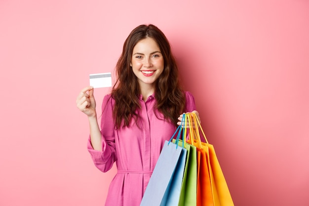 Mujer joven morena sosteniendo bolsas de la compra, mostrando tarjetas de crédito de plástico y sonriendo, de pie contra la pared rosa.