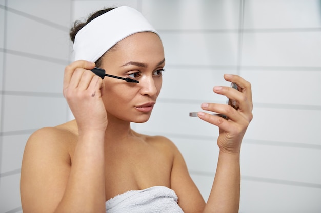 Mujer joven morena sosteniendo un espejo cosmético por un lado y rímel por el otro. Mujer aplicando rímel en sus pestañas y mirando a un espejo.