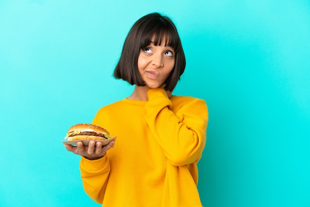 Mujer joven morena sosteniendo una hamburguesa sobre antecedentes aislados y mirando hacia arriba