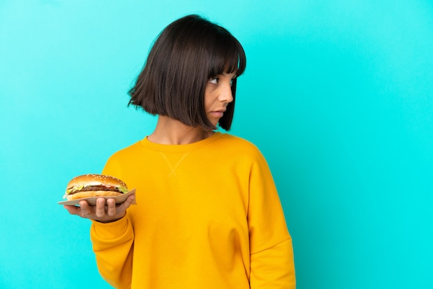 Foto mujer joven morena sosteniendo una hamburguesa sobre antecedentes aislados mirando hacia el lado