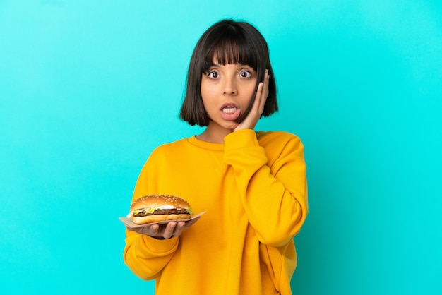 Mujer joven morena sosteniendo una hamburguesa sobre antecedentes aislados con sorpresa y expresión facial conmocionada