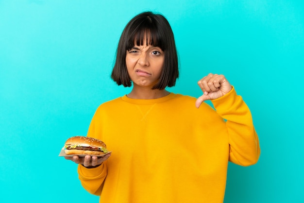 Mujer joven morena sosteniendo una hamburguesa sobre fondo aislado mostrando el pulgar hacia abajo con expresión negativa