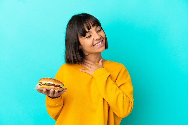 Mujer joven morena sosteniendo una hamburguesa sobre pared aislada mirando hacia arriba mientras sonríe