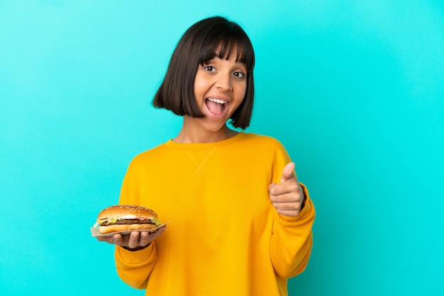 Mujer joven morena sosteniendo una hamburguesa sobre una superficie aislada con los pulgares hacia arriba porque algo bueno ha sucedido