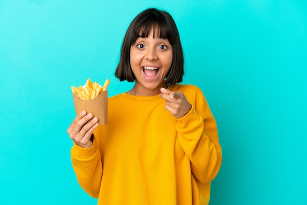 Mujer joven morena sosteniendo patatas fritas sobre fondo azul aislado sorprendido y apuntando hacia el frente