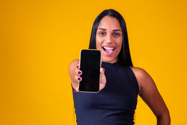 Mujer joven morena sosteniendo un teléfono celular con pantalla en blanco para texto sobre fondo amarillo