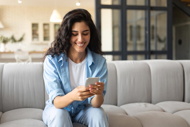 Mujer joven morena usando el teléfono celular en el sofá revisando el correo electrónico con video chat en línea en copia de casa