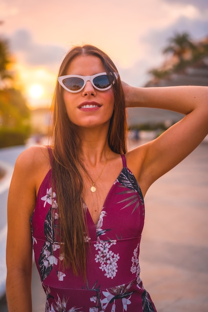 Mujer joven morena con un vestido floral marrón y gafas de sol modernas disfrutando del verano en la hora dorada